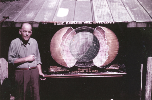 Unidentified man standing next to a Koreshan Unity poster showing "The Earth we inhabit".