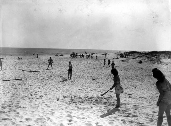 People playing softball on the beach with other activities in the background.