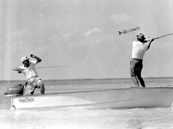 A fisherman reels in a barracuda as another fisherman takes caution :