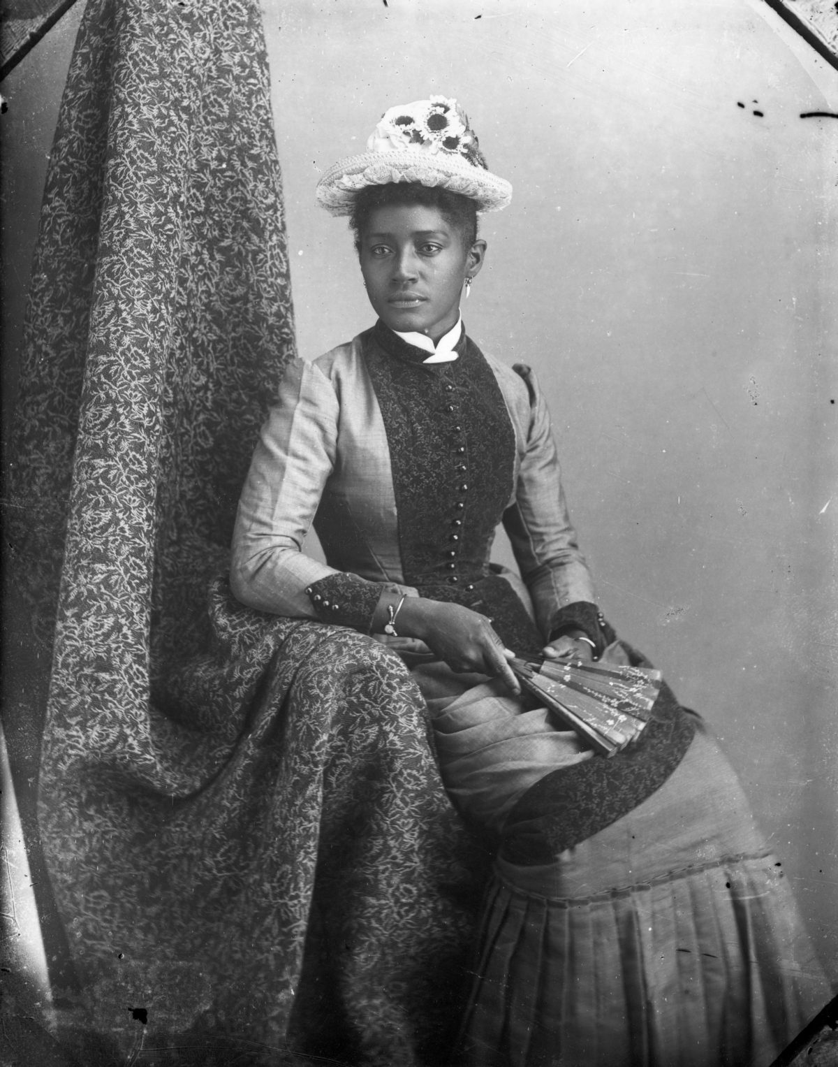 Seated woman wearing sunflower hat, holding a fan.