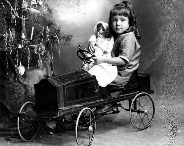 Unidentified girl holding her doll while sitting in a pedal car next to a Christmas tree - Pensacola, Florida.