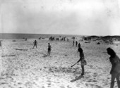 People playing softball on the beach with other activities in the background.