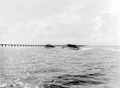 Two small yachts glide through the water with a pier in the background