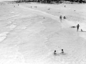 Children wading in the water with people watching from the beach.