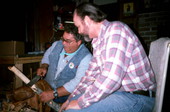 Teacher Bobby Johns showing apprentice Randy Stewart how to strip the bark off a white oak tree