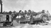Steam shovel filling ore cars pulled by narrow gauge steam engine