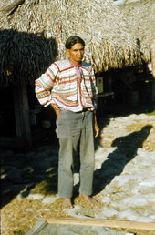 Unidentified Seminole Indian man standing by grass hut at the Dania Seminole Indian Reservation