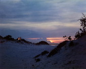 Scenic beach view showing people gathered around a bonfire in the distance during sunset.